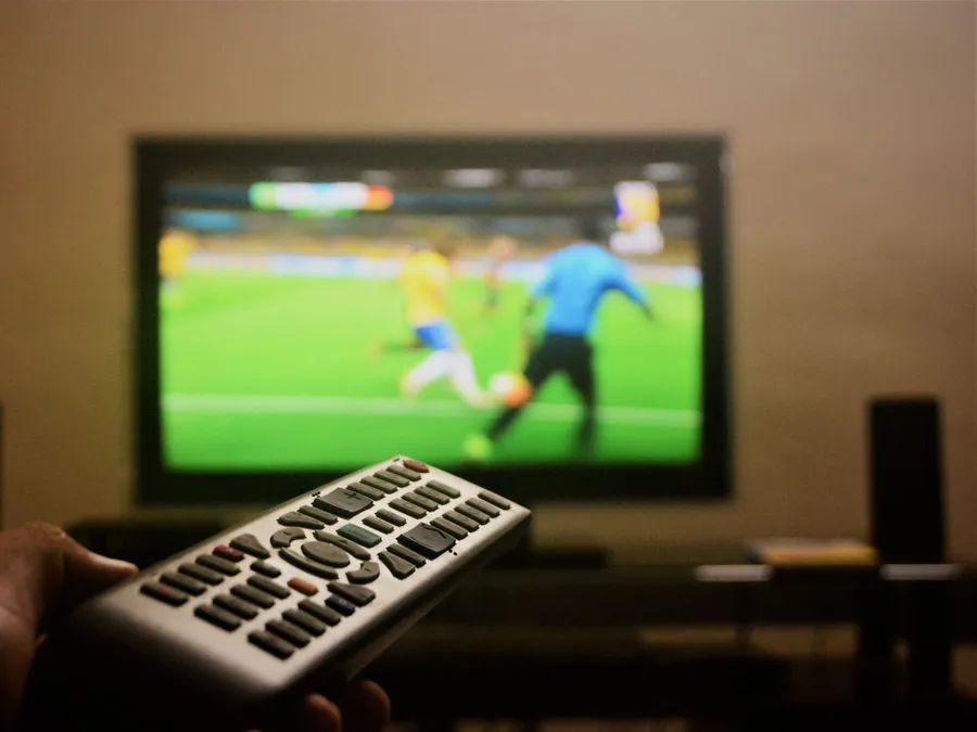 Hand pointing a remote control at a TV showing a fast-moving soccer match in a dim room